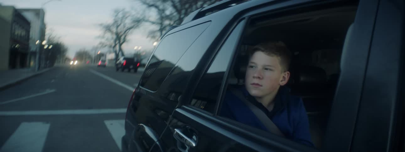 A boy sitting in the backseat of a vehicle looks out the window as it passes through a city