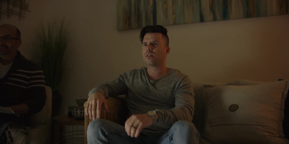 A white man sits on a couch in a living room with a relative next to him