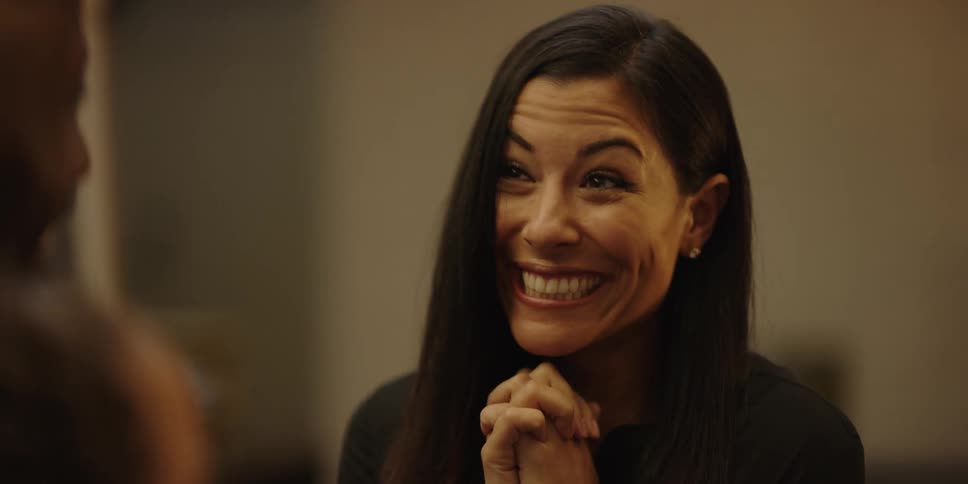 A woman with hands folded in front of her face brings them down and smiles in an excited and happy fashion as she looks at her family