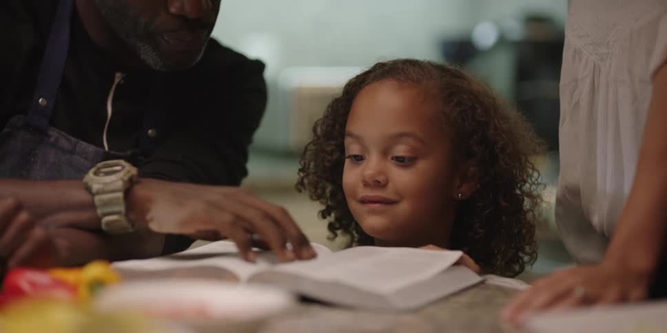 A father flips through pages of a book with his daughter as his wife watches on