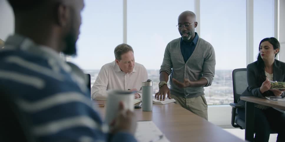 Four people in an office talk and look at a book while a woman also eats