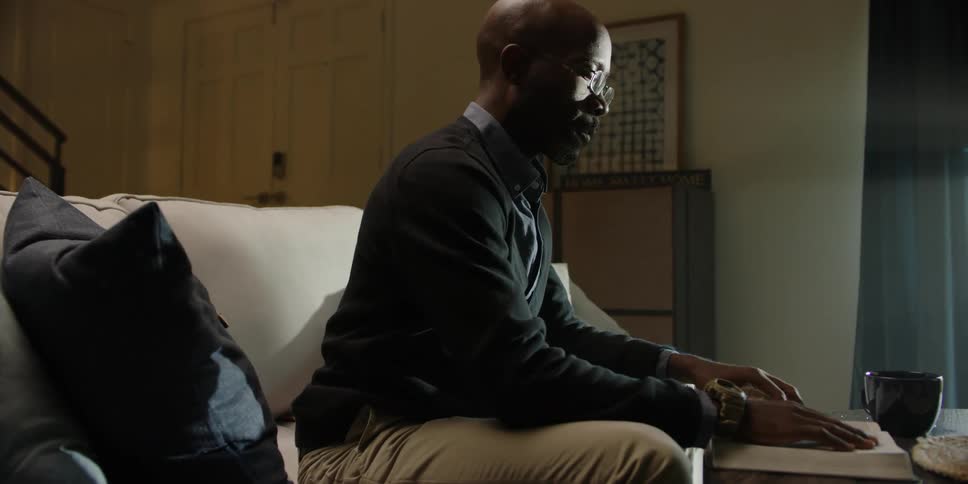 A blind man leans forward from couch to read a book on a table with his fingers
