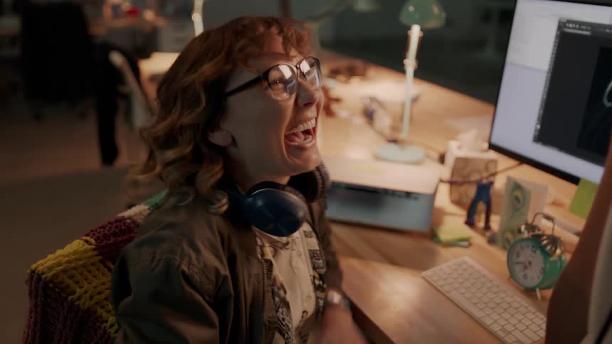 Two women sitting by a desk talking with a computer and laptop in the background