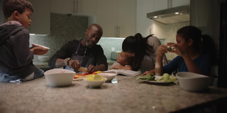 A black family sits around a counter top together talking and preparing food