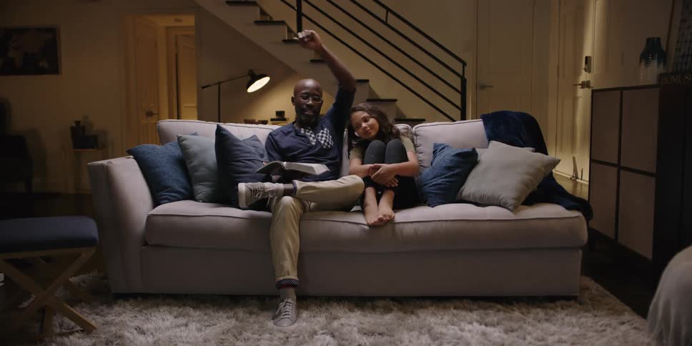 A girl twirls and dances over to sit on the couch next to her dad who puts his arm around her and reads