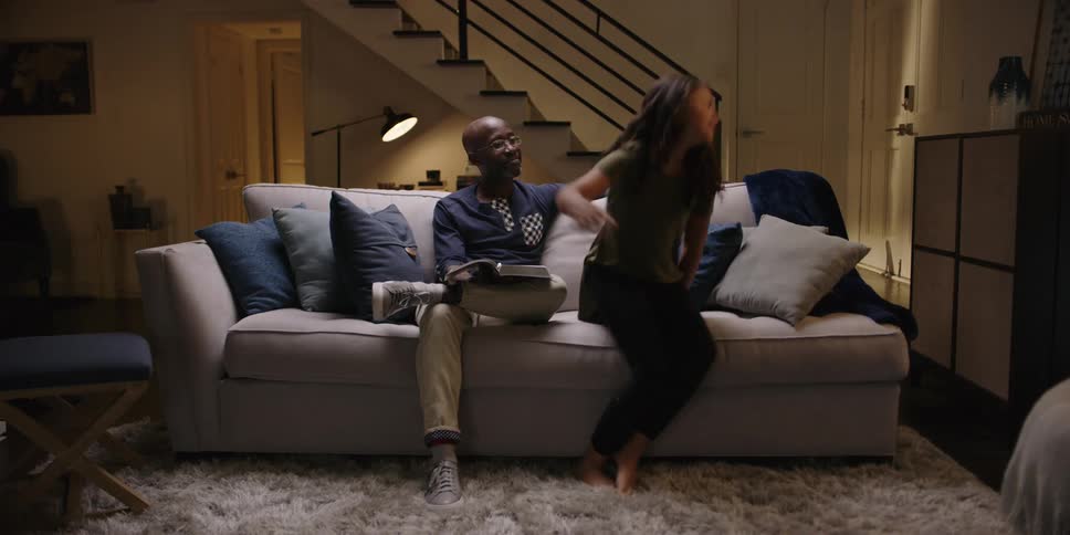 A girl sitting next to her dad while he reads gets up from the sofa and walks upstairs