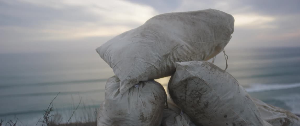 Stacked filled bags rest on ground near ocean and promontory as sun reflects from water