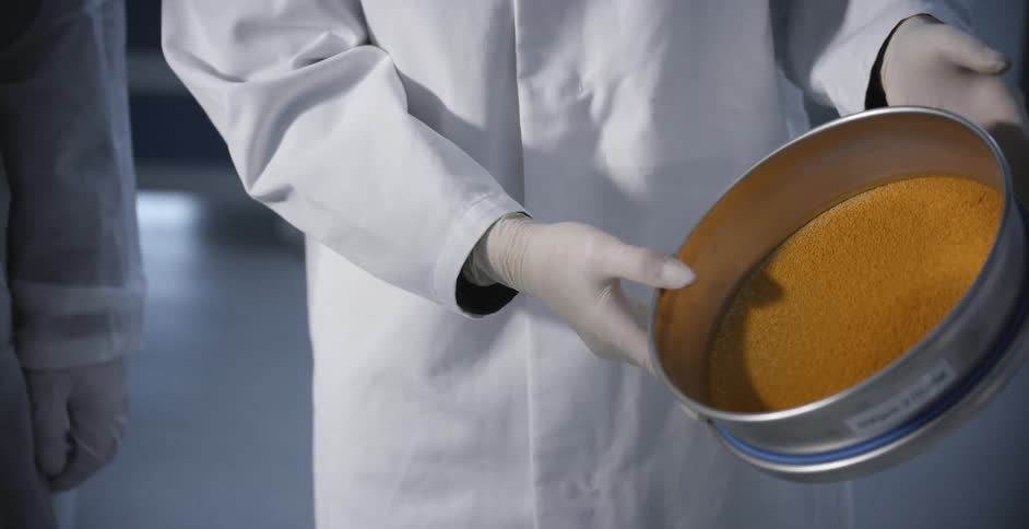 A scientist in a lab holds and tilts a canister with orange particles in it