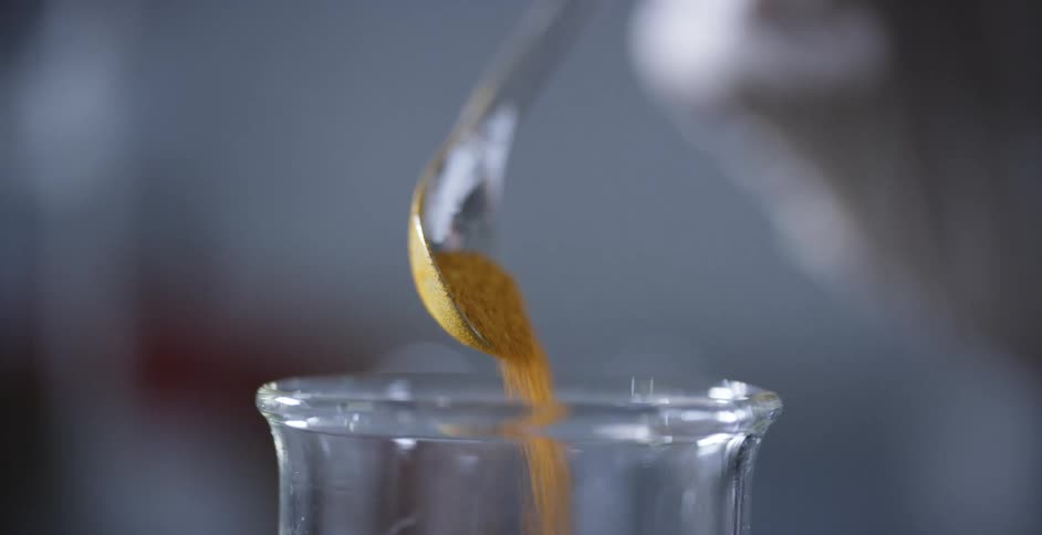 A person in a lab pours orange granules in a glass flask with a spoon