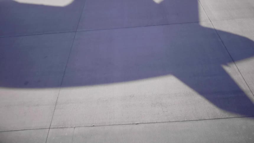 The shadow of a small aircraft is shown taking off from a concrete airfield