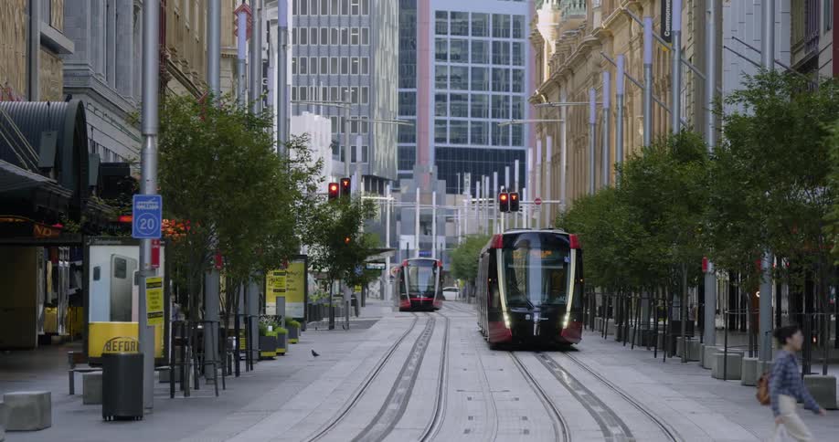Two city trains sit on the tracks while a pedestrain crosses in front