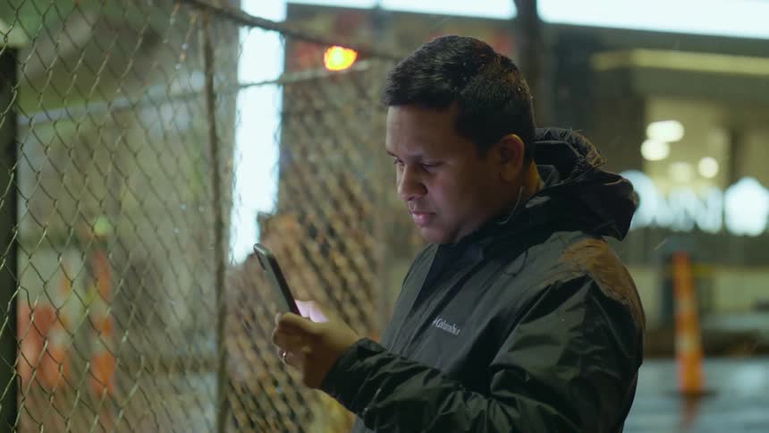 A man stands on a city street with snow falling and looks at his cell phone