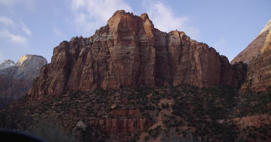 Traveling past mountain and escarpment dotted with scrub under slightly cloudy blue sky