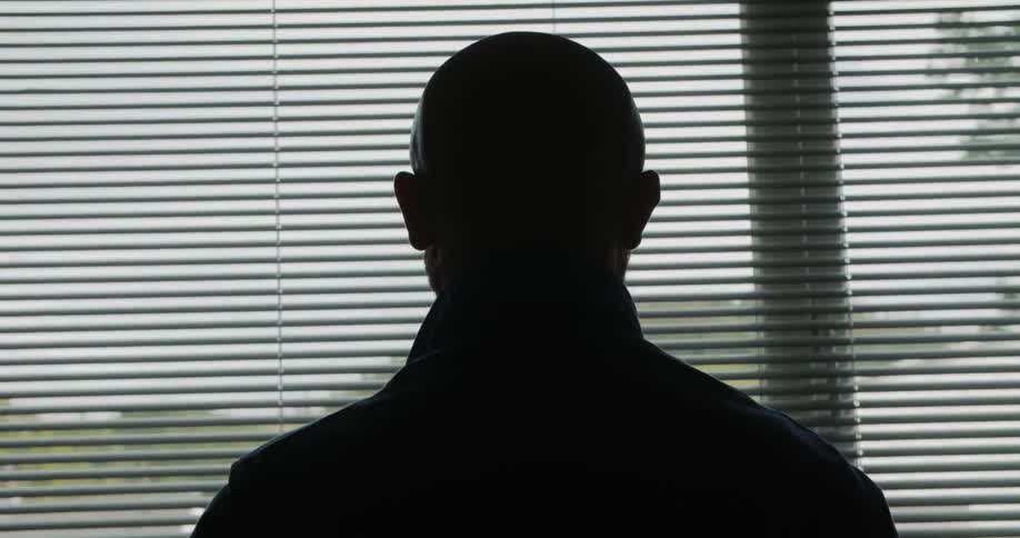 Bald man cast into silhouette by incoming light stands looking out window through open blinds