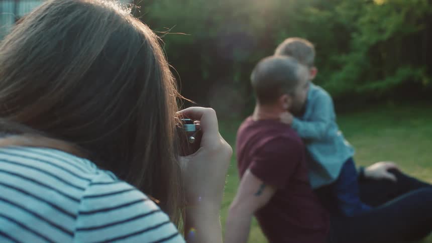 Woman takes a photograph of father and son sitting on the grass in the garden
