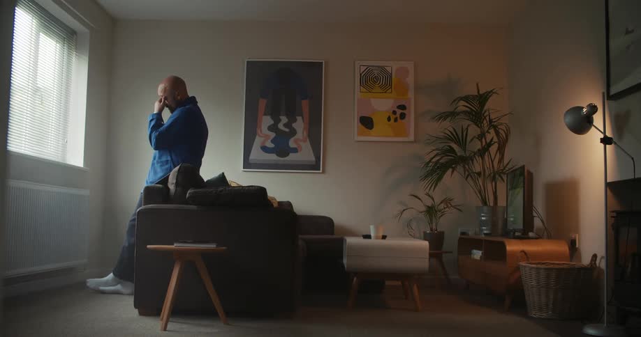 A man leans against his couch as he looks out of his window and then takes off his glasses and rubs his eyes