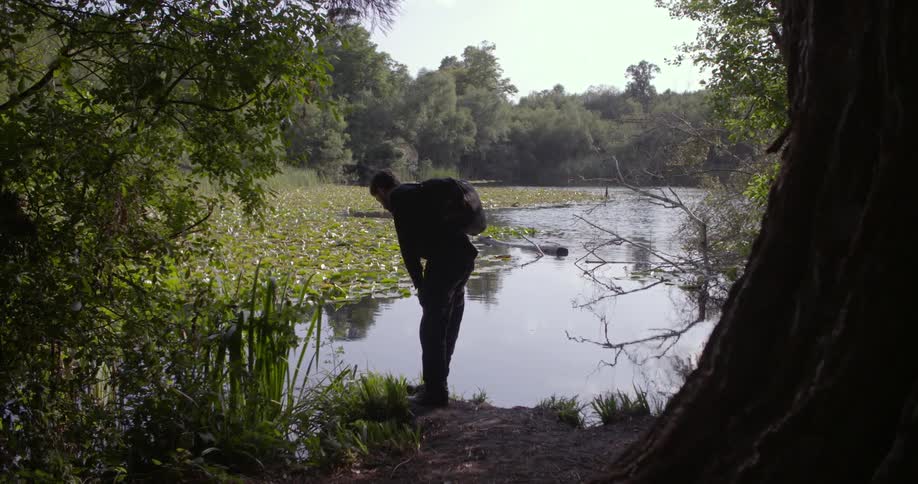 A man wearing a backpack stands up and continues to look down at the pond water
