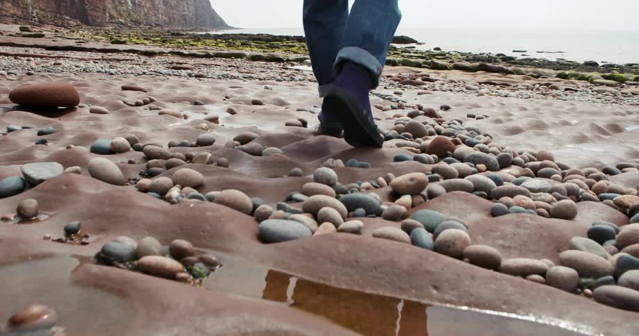 Person walks across stone covered terrain near cliff toward water