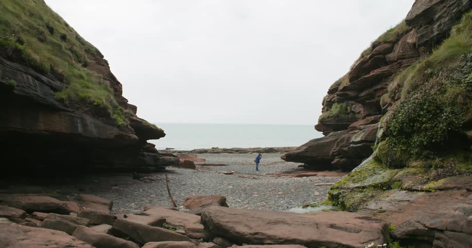A person walks along a gravel beach by tall mossy hills