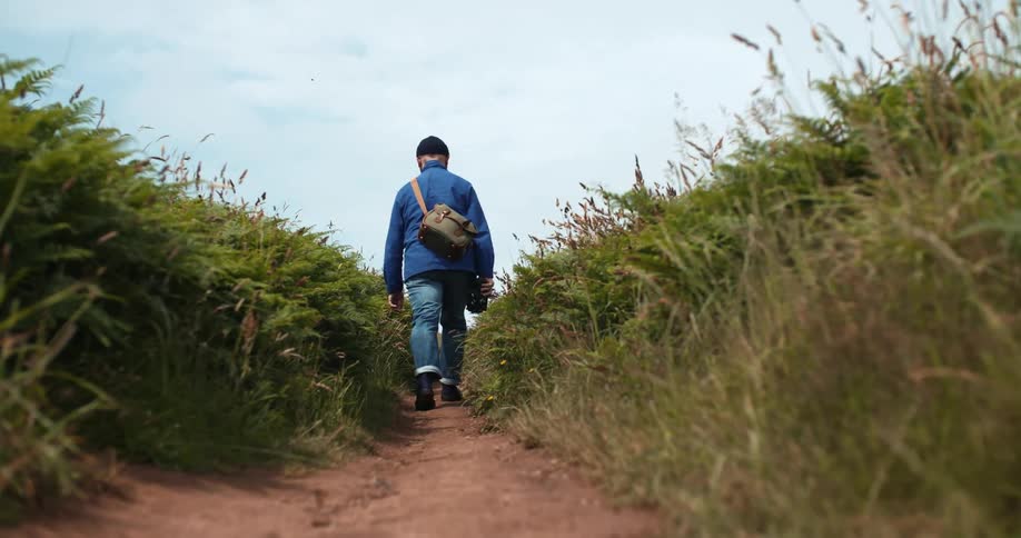 A man carrying a video camera and a bag walks on a narrow dirt trail between tall grass on a sunny day