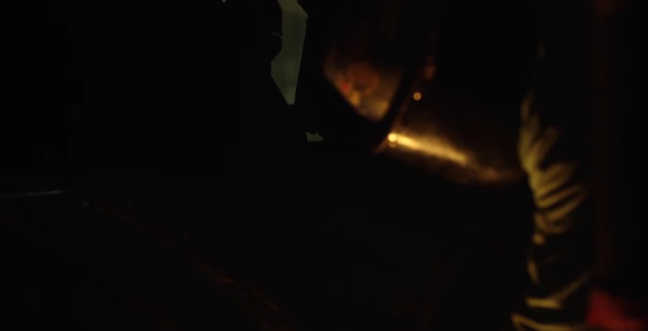 A man wearing gloves a helmet and a mask kneels outdoors by a campfire