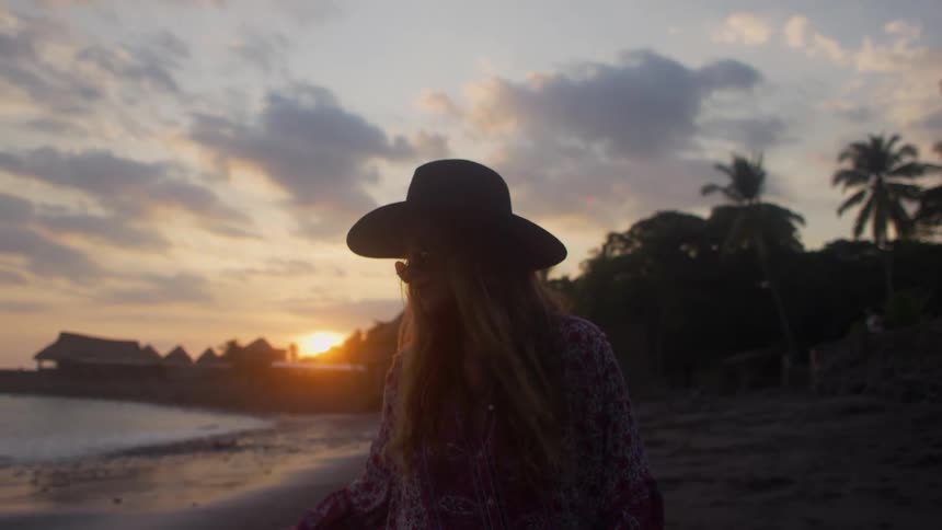 Woman wearing a wide hat walks on the beach at sunset next to the sea