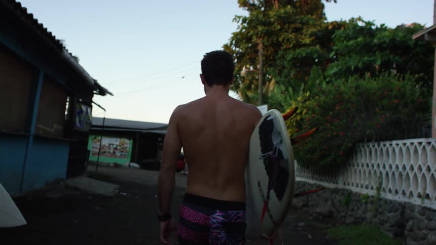 A white male carrying a surfboard walks on a dirt path near buildings