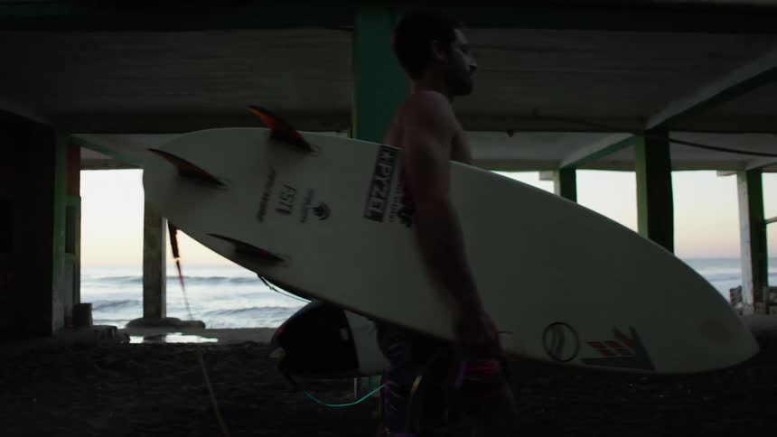 Three friends carry their surfboards on the sand underneath the pier as ocean waves come to shore