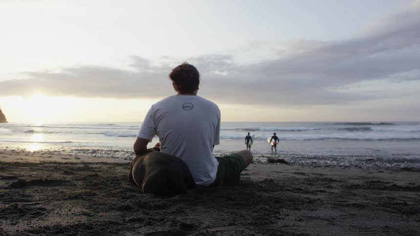 A man sits on the beach with his dog while watching two surfers walk in to the water