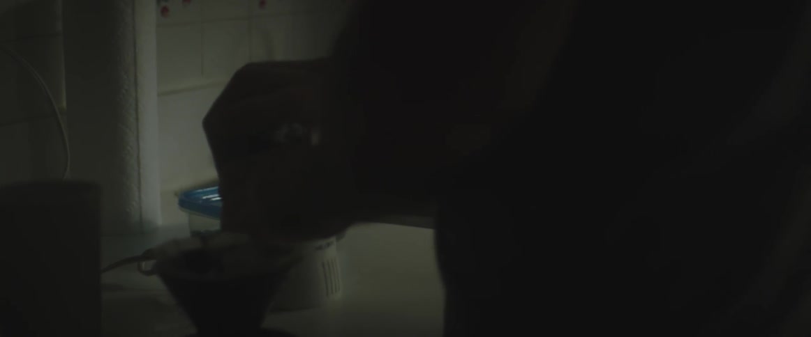 A man standing in a dimly lit kitchen uses a coffee grinder