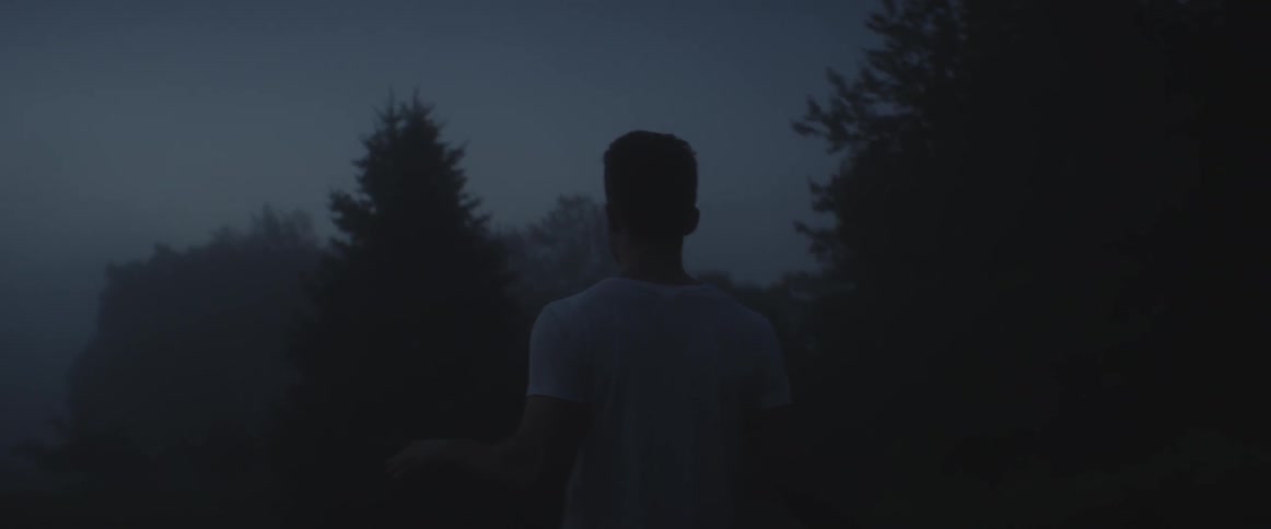 A man in a t-shirt walks through a foggy, forest at night