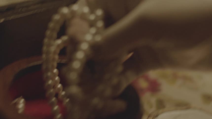A woman's gloved hand reaches into a box to pick up a strand of pearls