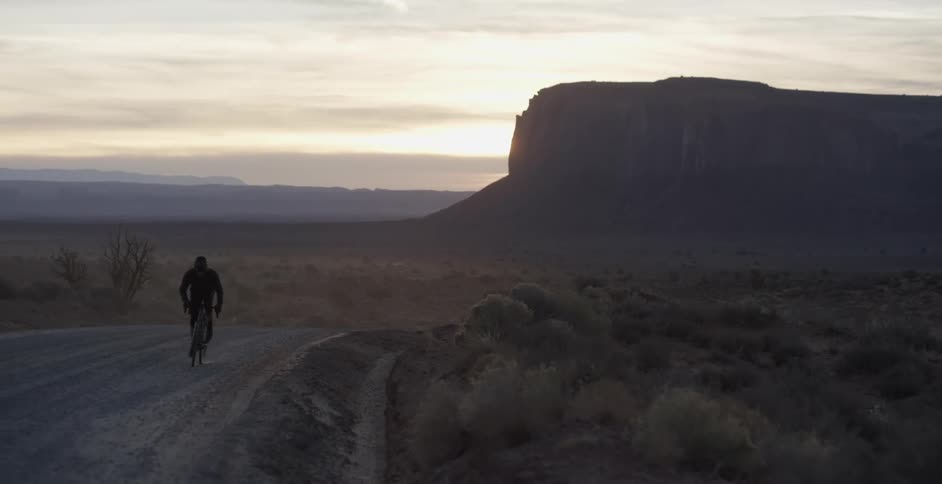 A man pedals hard riding a bike on a dirt road with rock formations behind at sunrise