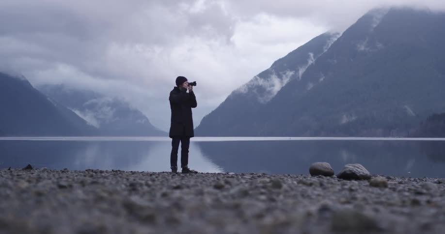 A man holding a camera with a lake and mountains in the background