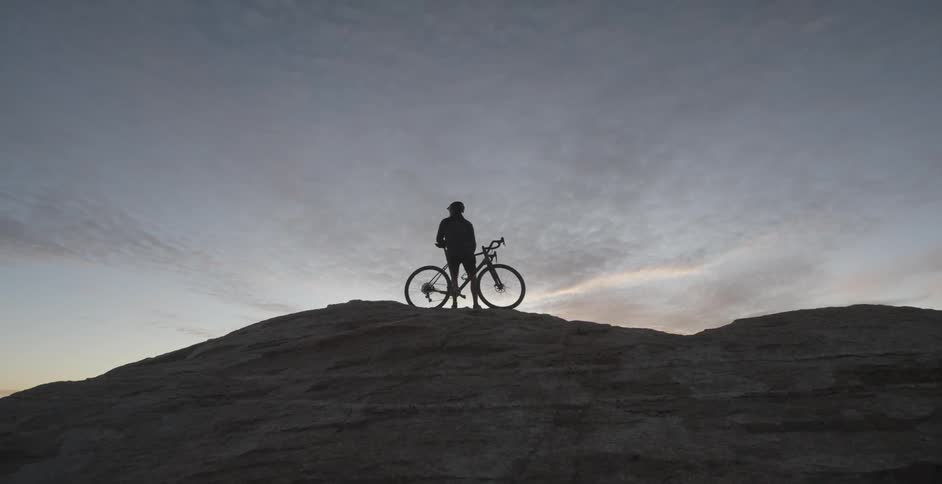 A cyclist stands on a ridge with their bike looking out at the sunset sky