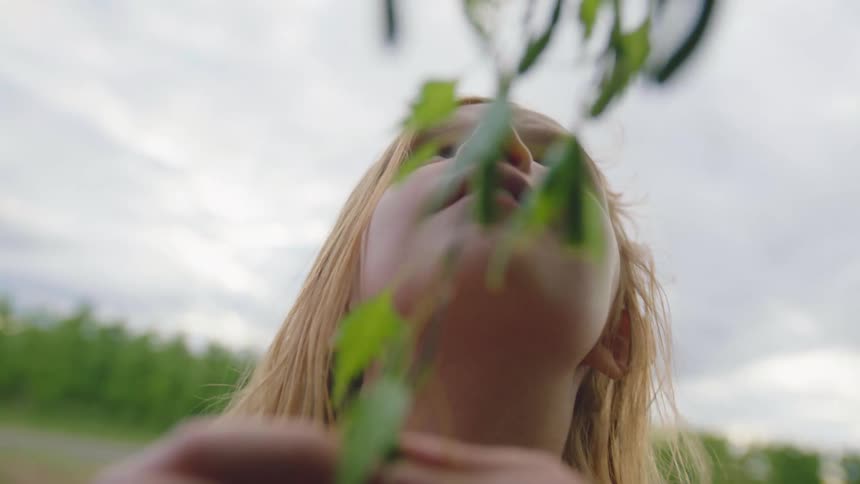 A young white female teen stands near a tree and plays with the leaves on a branch