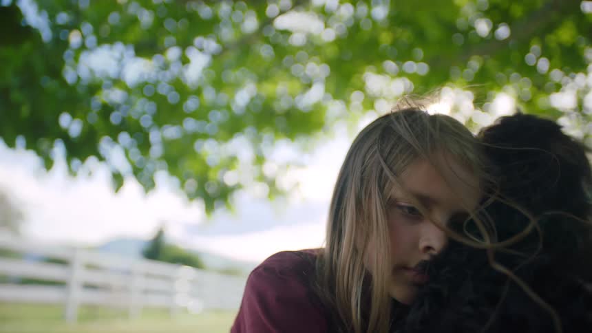 Blonde little girl cuddles a dark dog under the tree