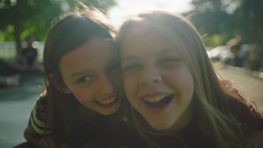 Two girls laugh and play on a trampoline as other kids play on the ground behind them