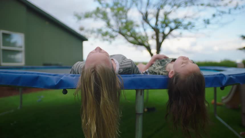 Two white girls recline on a trampoline in the yard of a house