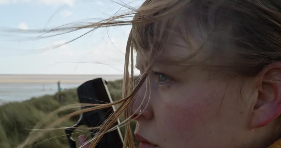 A white woman stands on a coast and raises her hand to reveal a gun