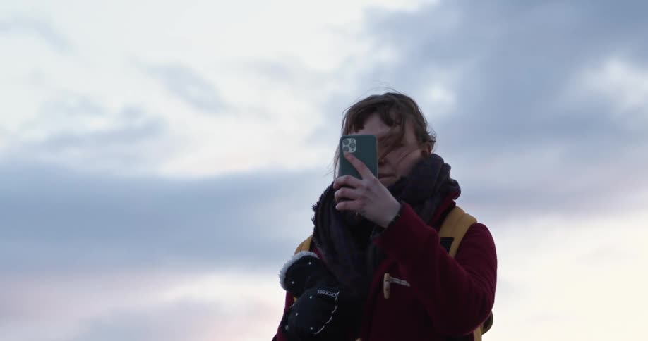 A woman wearing a big scarf and coat holds her phone up to her face to take a selfie outside