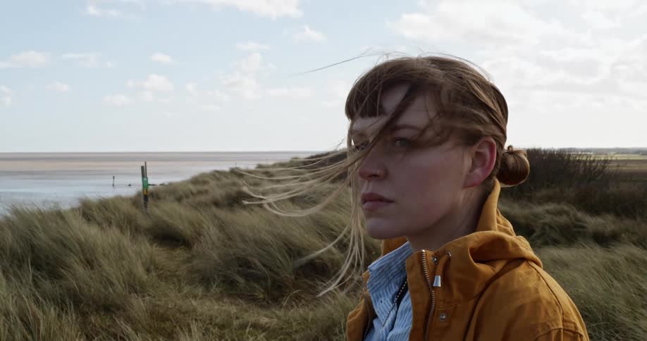 A white woman stands near a coastline and looks into the distance
