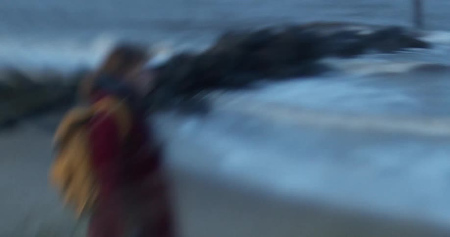 A person wearing a coat and backpack stands on the beach looking out at the waves crashing against the rocks