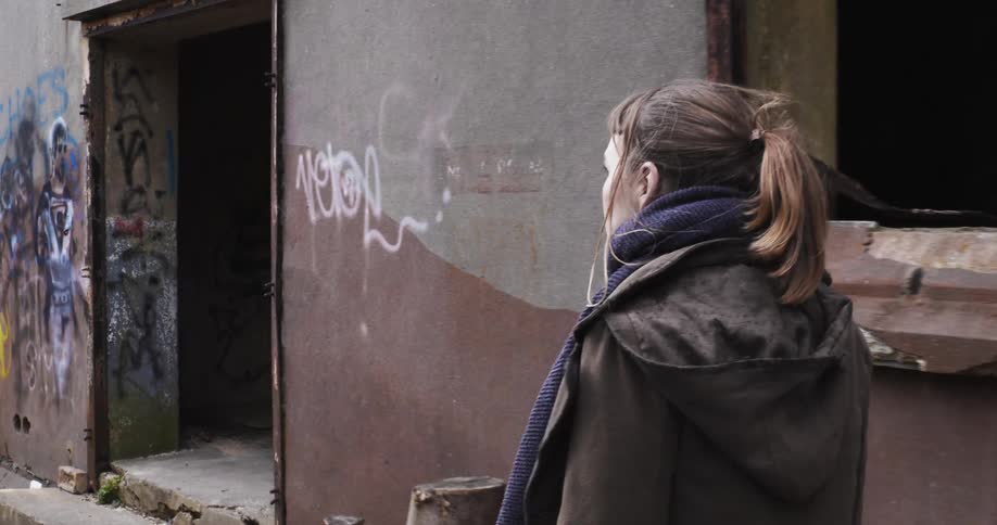 A white woman strolls down a sidewalk and turns to enter a decrepit urban building