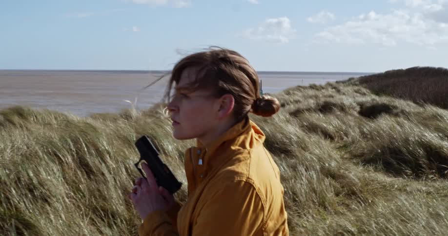 Woman carries a handgun while walking on a grass field near the ocean