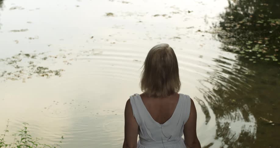 A white woman in a dress stands next to a lake