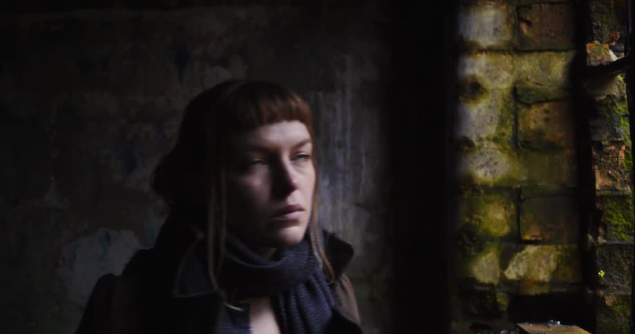 A woman looks out an opening in an old stone building before turning to walk away