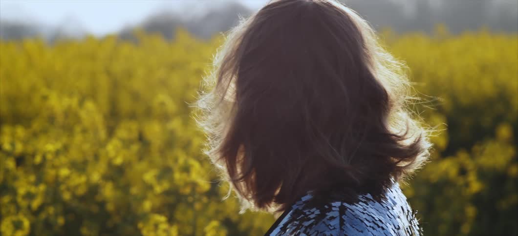 A white woman stands in a field of yellow flowers as the wind blows her hair