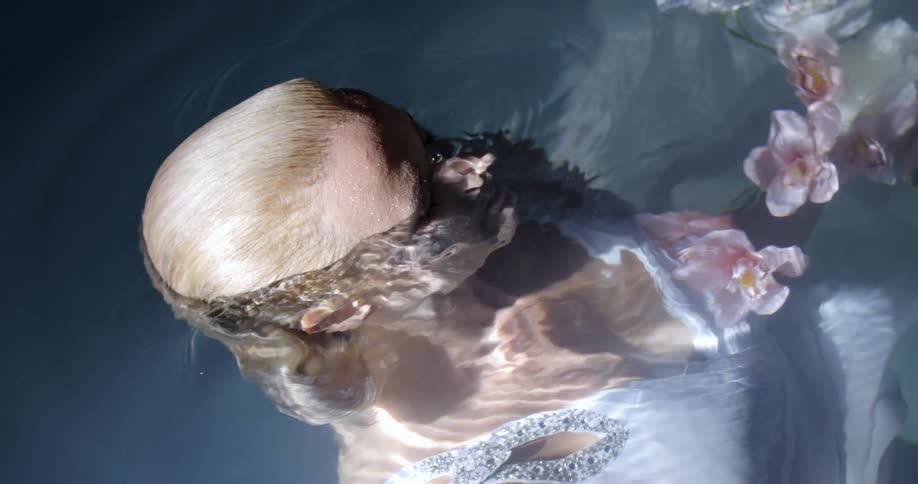 A blonde white woman in a dress slowly submerges herself underwater