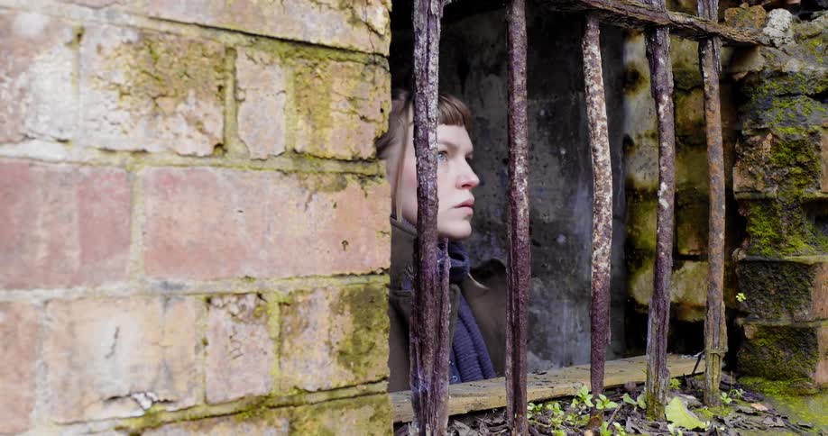 A woman approaching a barred mossy window from the inside of a ruined building and gazing outside